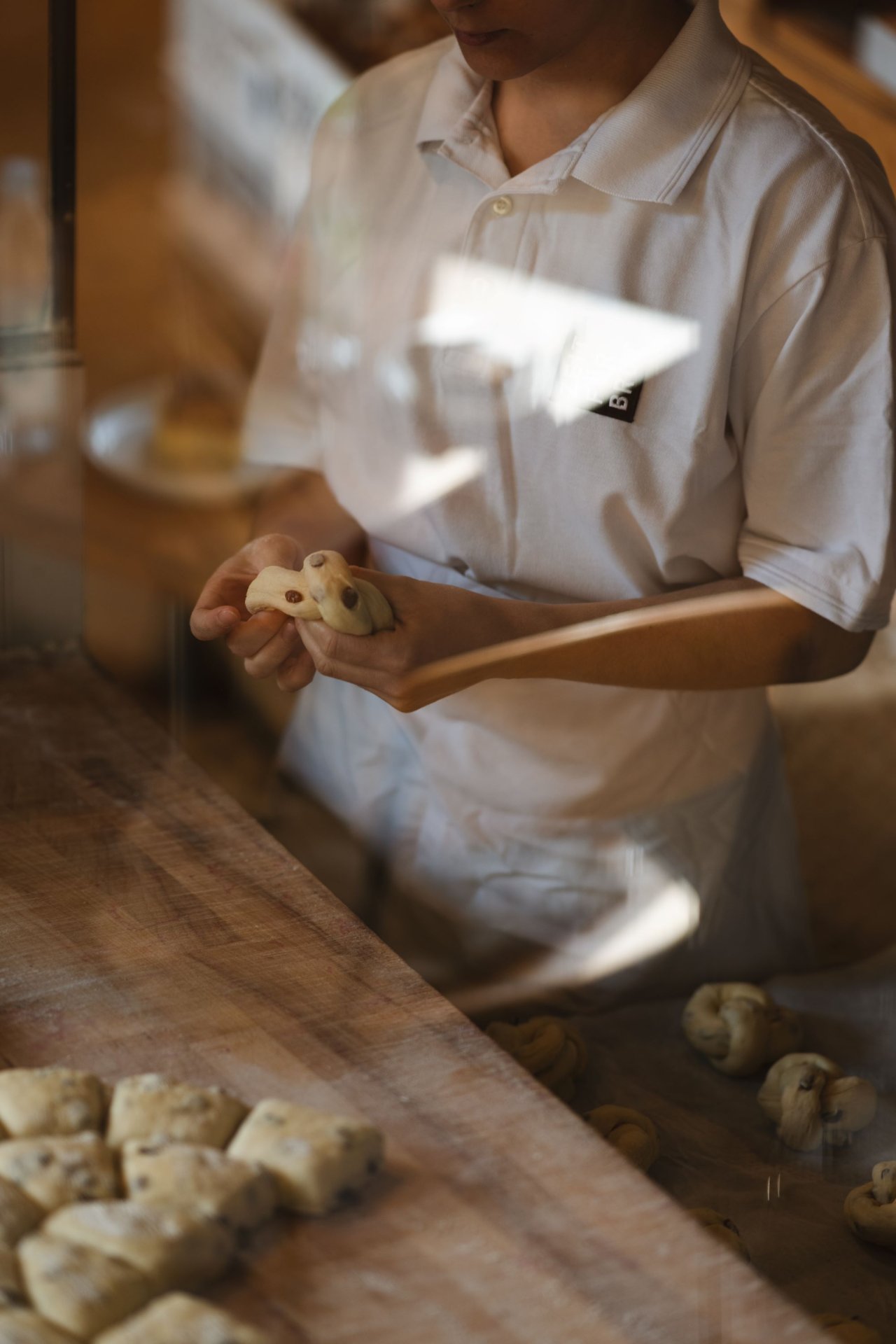 Eine Person in einem weißen T-Shirt formt Teig mit beiden Händen. Der Hintergrund zeigt eine Holzoberfläche und weitere Teigstücke.