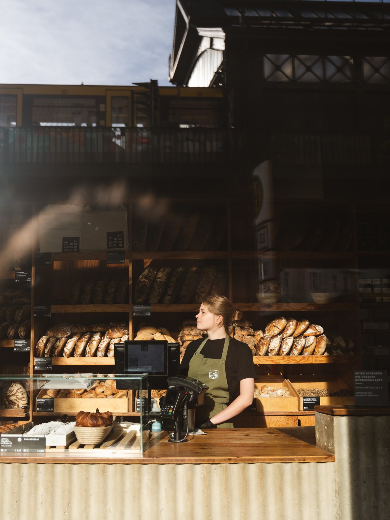Bäckerei mit Holzregalen voller Brote. Eine Person steht hinter der Theke, trägt eine grüne Schürze und hat die Hände auf der Theke.